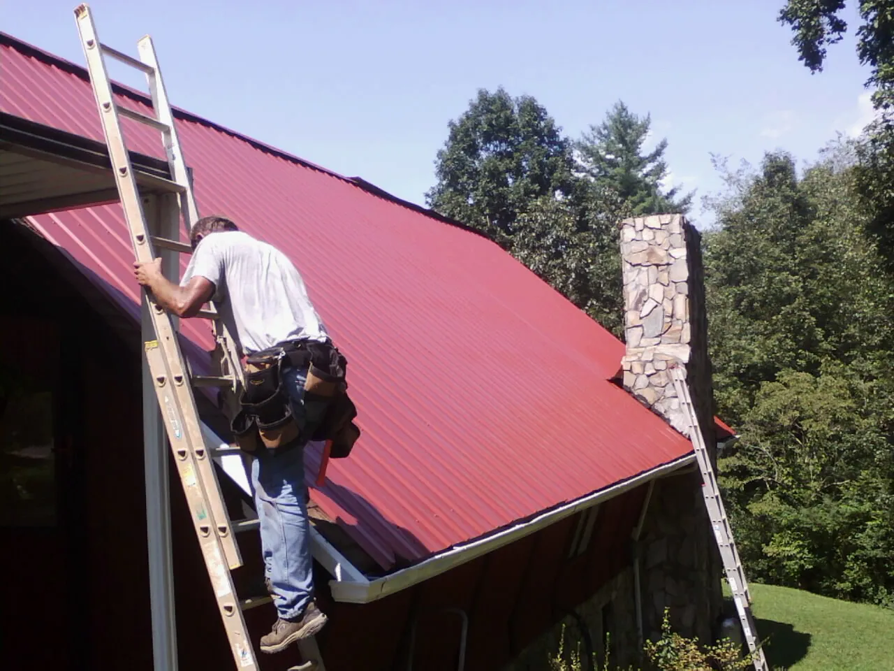 Roofer on a ladder beside a red metal roof and stone chimney — Soffit & Fascia Repair by Roofing Brothers in Woodville